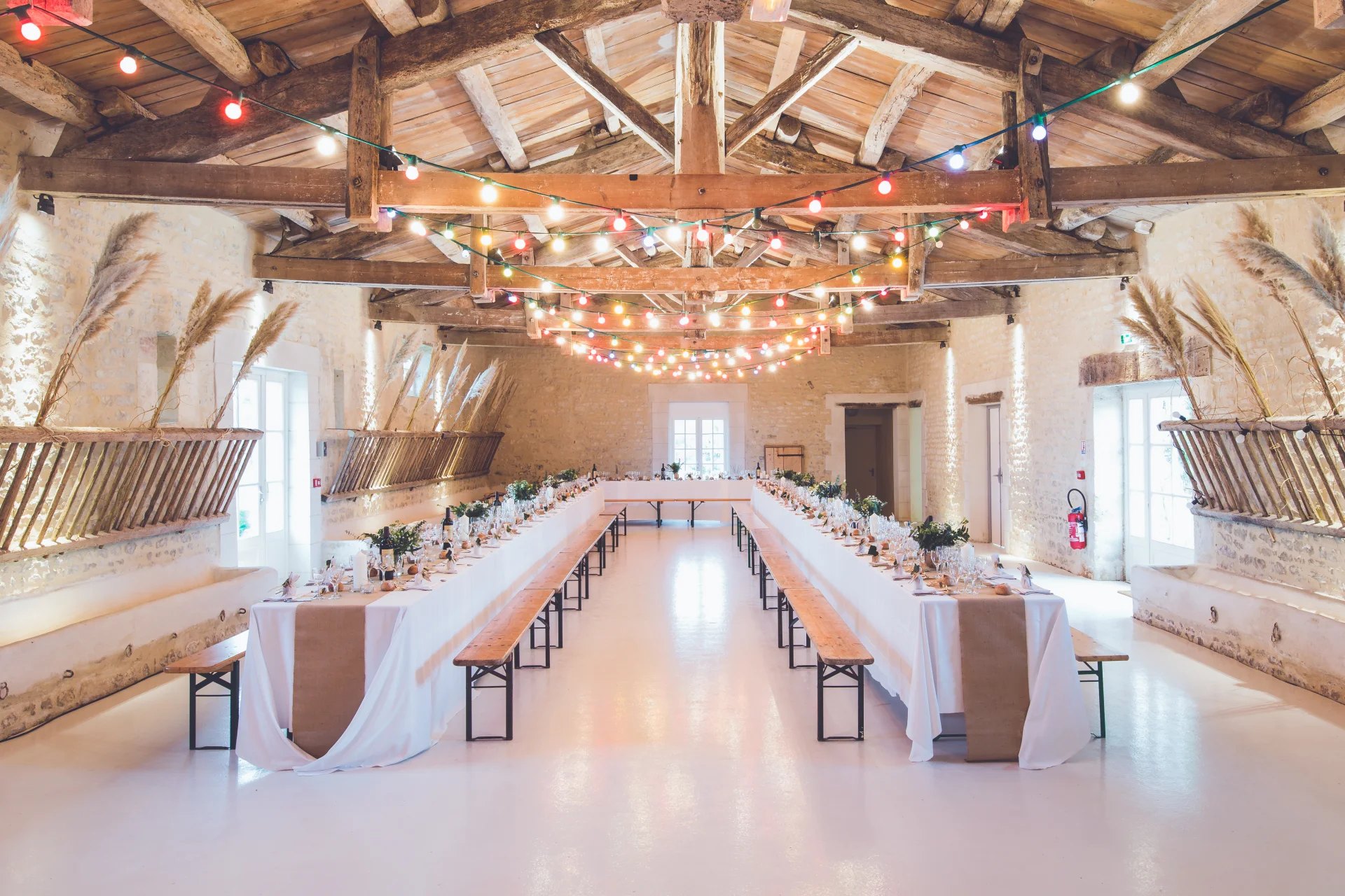 Fotografie eines Hochzeitssaals mit langen weiss gedeckten Tafeln, Holzbänken und Lichterketten unter sichtbaren Dachbalken